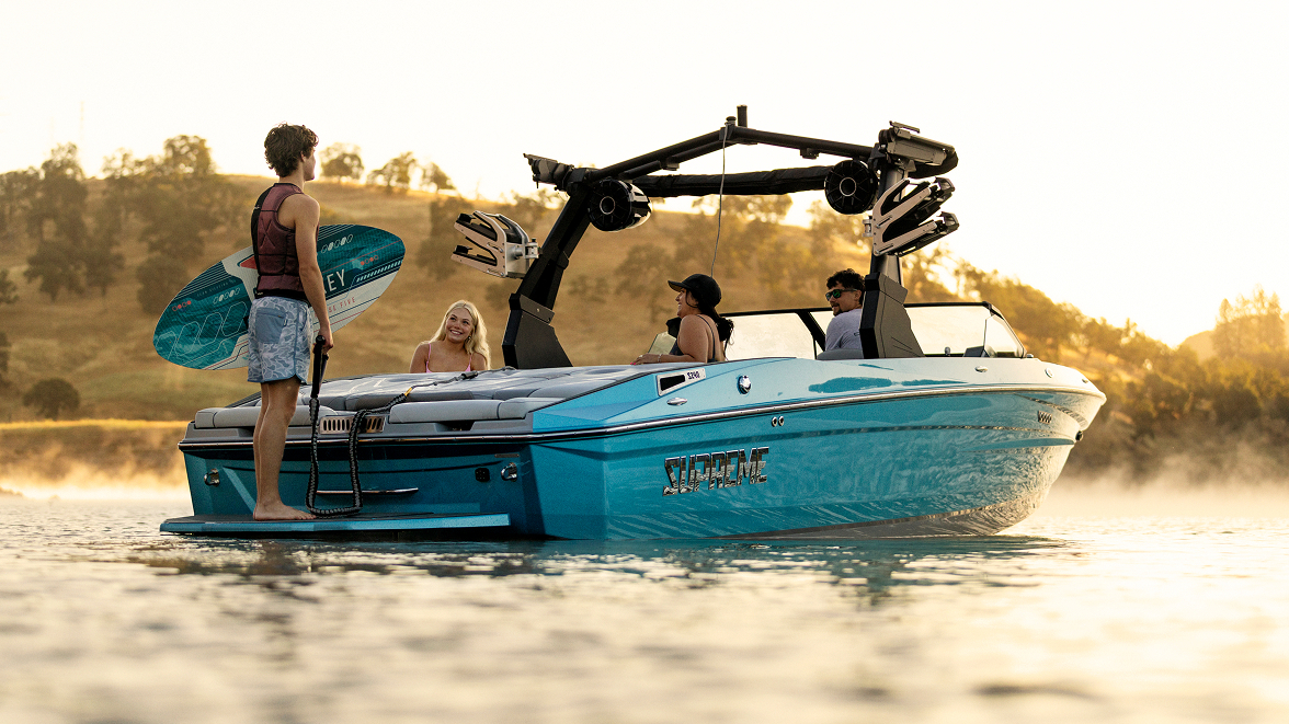 Four people are on a blue Supreme Boats S240 wakeboard boat in calm water at sunset. One person stands on a swim platform holding a wakeboard while the others sit and talk. Trees and hills are in the background.