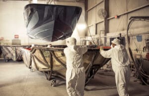 Two workers in protective suits guide the hull of a Supreme S220 boat being lifted by a crane inside an industrial workshop.