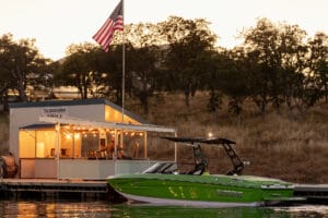 A Supreme S220 is moored by a small dockside restaurant with string lights, an American flag, and hilly terrain in the background during sunset.