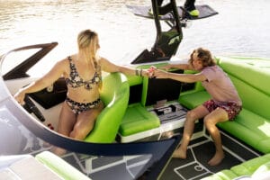 A woman in a floral bikini hands a drink to a seated young person in red shorts on the green-seated Supreme S220 boat, with someone wakeboarding in the background.