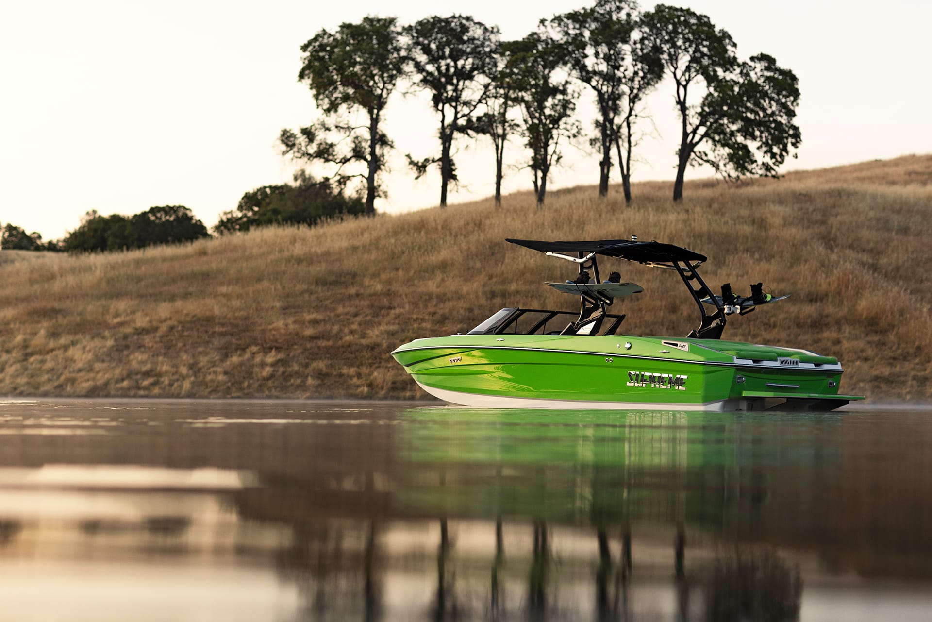A bright green Supreme S220 boat with a black canopy sits on calm water near a grassy hill with trees.