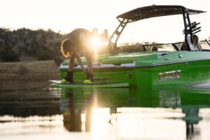 Person adjusting their wakeboard at the edge of a green Supreme S220 speedboat on a calm lake during sunset.