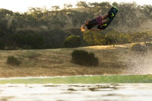 A person performing an airborne trick on a wakeboard over a body of water, with a rural landscape in the background, is effortlessly pulled by the sleek Supreme Towboats S220.