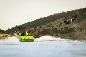 A person is wakeboarding behind a green Supreme Towboats S220 on a lake, with a hilly landscape in the background.