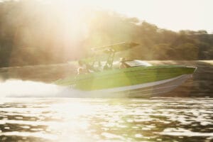 A Supreme S220 green motorboat speeds across the water with sunlight glaring in the background and a wooded shoreline visible.