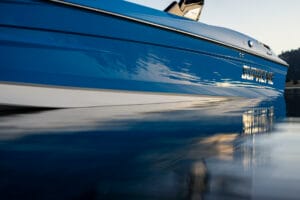 Close-up of a blue Supreme Boats S240 named "SUPREME" partially submerged in calm water with a blurred reflection of the boat on the water's surface.