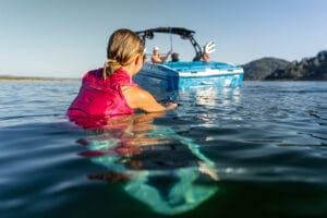 A person in a pink life jacket swims towards a Supreme Boats S240 with people on board in a calm body of water, with mountainous terrain in the background.