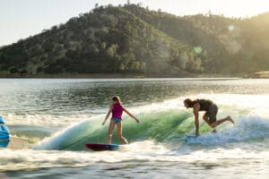 A girl and a man ride a wave on surfboards next to a Supreme Boats S240 in a lake, with a forested hillside in the background.