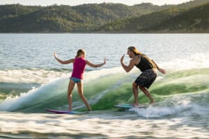 Two surfers, a man and a girl, ride waves side by side on a lake surrounded by hilly terrain. The girl reaches out with both hands while the man smiles and gestures with one hand, clearly enjoying the powerful wake generated by their Supreme S240.