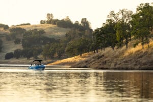 A blue Supreme Boats S240 floats on calm water near a shore lined with trees. The background features rolling hills with scattered trees under a pale sky.