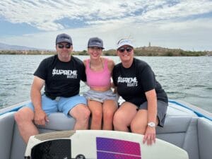 Three people, likely from the Gilmore family, wearing Supreme Boats hats sit together on a boat in open water. Two women in pink and black tops are flanked by a man in a black shirt and blue shorts. A wakeboard rests in front of them, hinting at their Lake Havasu adventure.