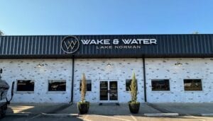 Front exterior view of Wake & Water Lake Norman building, featuring a white brick facade with large windows, a central entrance door, and two potted plants on either side—an inviting scene at this premier dealership for Supreme Boats and Centurion Boats.