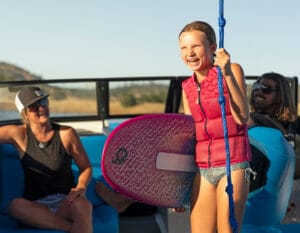 A smiling child in a red life jacket holds a pink surfboard on a boat, with two adults sitting behind her, experiencing The Supreme Life with Supreme Boats.