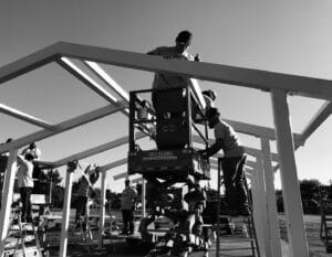 Several individuals working together to construct the frame of a building using ladders and a scissor lift on a clear day, truly experiencing The Supreme Life.