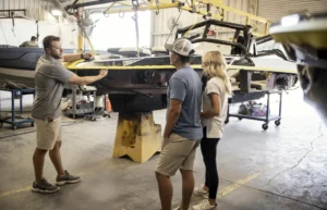 A man holds a tape measure against a boat in a workshop, explaining something to a man and a woman about their Supreme Life on the water.