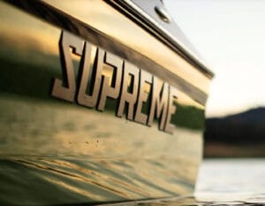 Close-up of a boat's side with the name "SUPREME WAKEBOATS" prominently displayed in large letters, reflecting sunlight off the water surface. The background shows a blurred landscape with calm water.