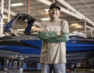 A man stands with arms crossed in front of a blue Supreme Wakeboat inside a warehouse, wearing a tan shirt over a green long-sleeve shirt and a cap.