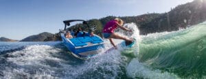 A person in a pink shirt wake surfing behind a blue boat on a lake, with two people seated in the boat enjoying the moment. The shoreline is visible in the background. Could this be the homepage image of 2025?