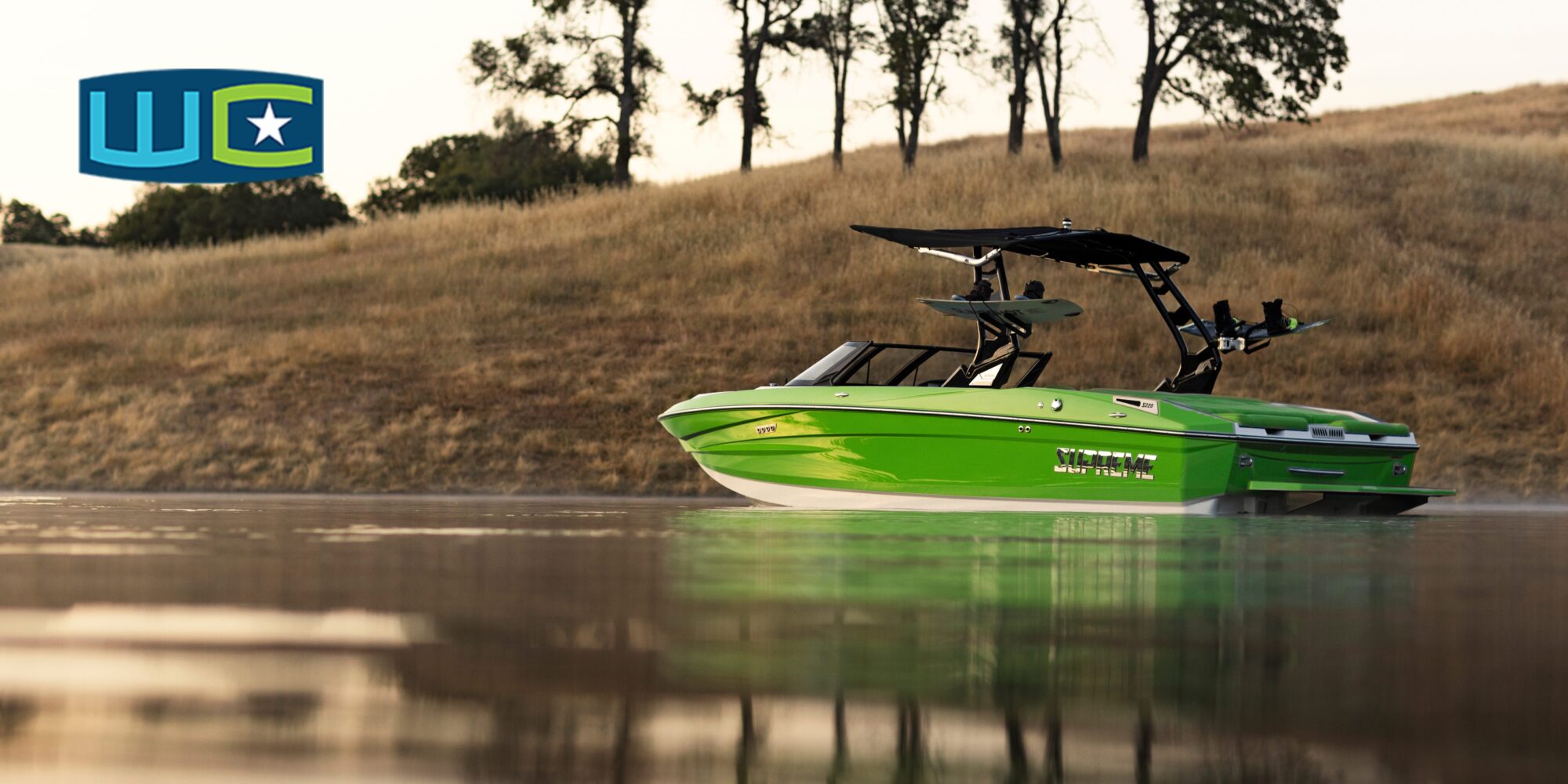 A bright green motorboat with a black canopy floats on calm water near a grassy shoreline with trees in the background. A logo with letters 