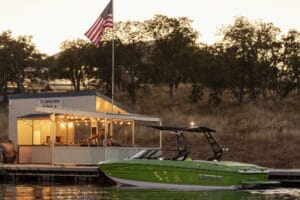 A green boat is docked near a small building adorned with string lights and an American flag, set against a backdrop of trees and a sunset sky—a serene preview to the 2025 Supreme Boat Show extravaganza.