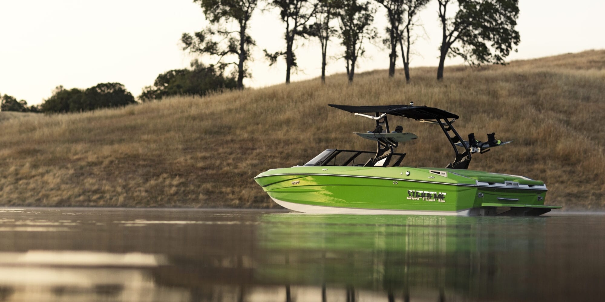A bright green motorboat, possibly a Supreme Boat with a wakeboard tower, floats on calm water, reflecting the landscape. Distant trees are visible on a grassy hill in the background.