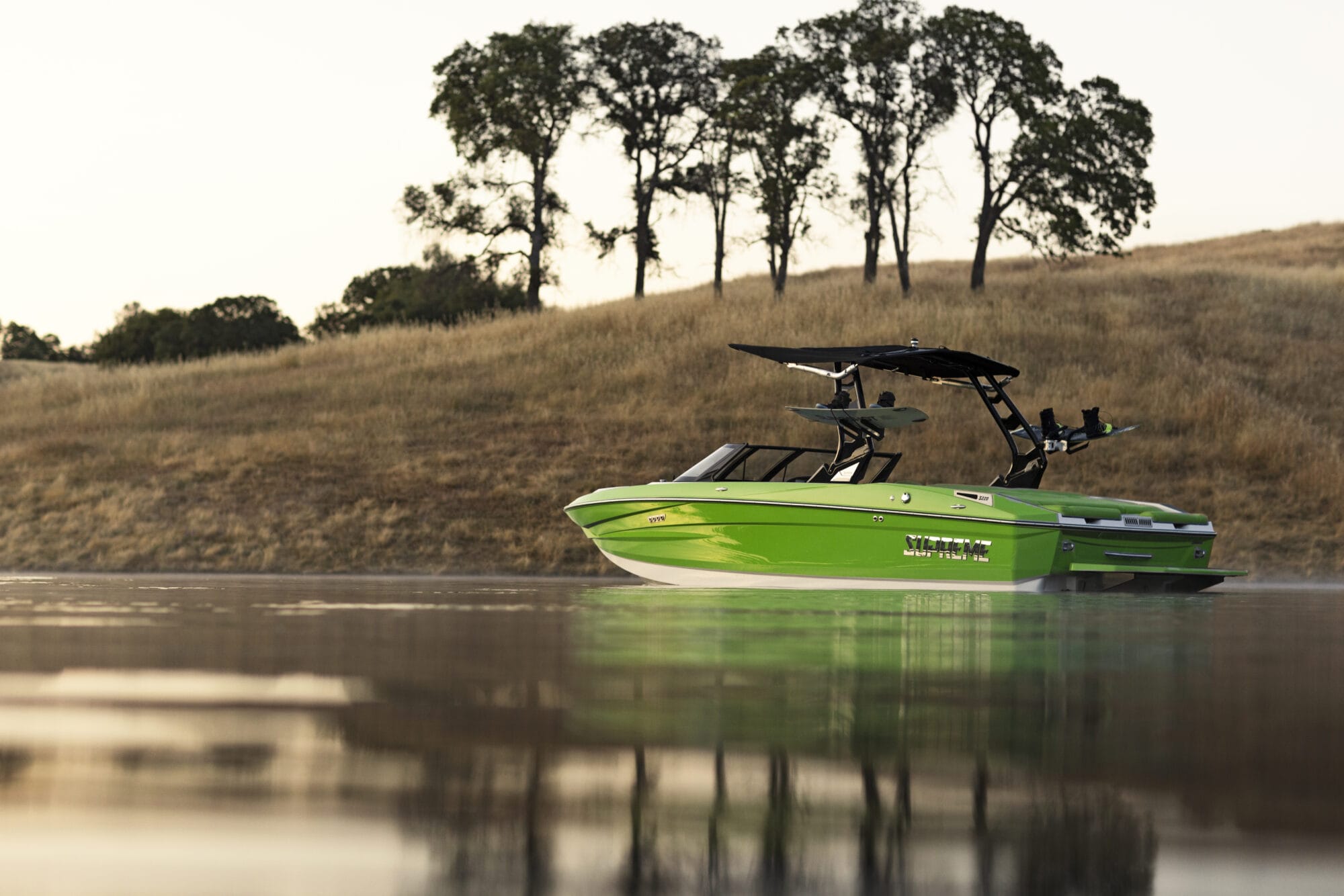 A bright green motorboat, possibly a Supreme Boat with a wakeboard tower, floats on calm water, reflecting the landscape. Distant trees are visible on a grassy hill in the background.