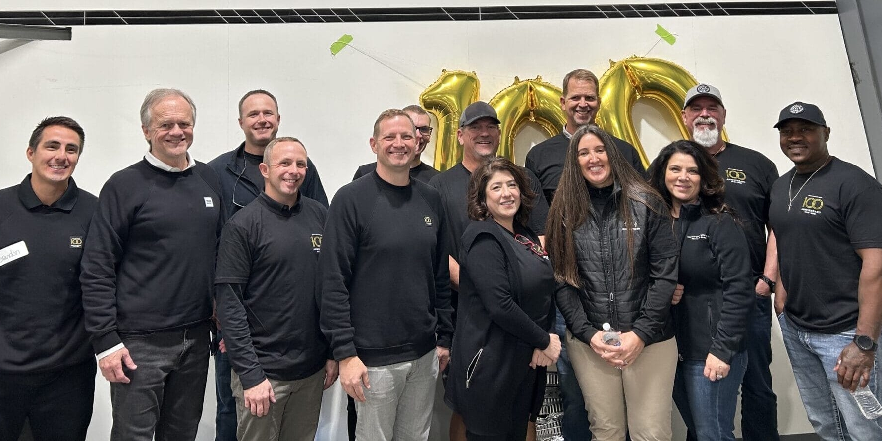 A group of people stands indoors in front of large gold balloon numbers and silver wall panels, all proudly wearing black shirts emblazoned with the Centurion logo, smiling at the camera.