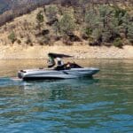 A speedboat with a black canopy cruises on a calm lake near a forested hillside.