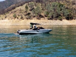 A speedboat with a black canopy cruises on a calm lake near a forested hillside.
