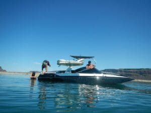 Three people enjoy a sunny day on a lake with a speedboat. One person is diving into the water, while the other two sit on the boat. Rocky hills are visible in the background.