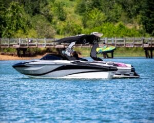 A sleek black and white motorboat with a wakeboard attached, cruising on a calm lake near a wooden pier, surrounded by green foliage.