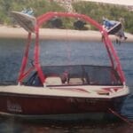 Red and white wakeboarding boat anchored on a lake, featuring a tower with speakers and wakeboards. Shoreline with greenery in the background.