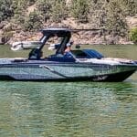A modern motorboat with people inside navigates on a calm, greenish lake with a rocky, tree-lined shoreline in the background.