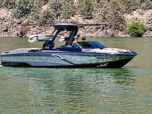 A modern motorboat with people inside navigates on a calm, greenish lake with a rocky, tree-lined shoreline in the background.