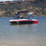 A red and white speedboat with a black canopy is on a lake, near a rocky shoreline. Wakeboards are mounted on the boat.