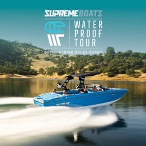 A blue Supreme boat speeds across a lake with passengers onboard, showcasing the thrill of the "Supreme Wakeboats Waterproof Tour" Drive-N-Ride Sales Event.