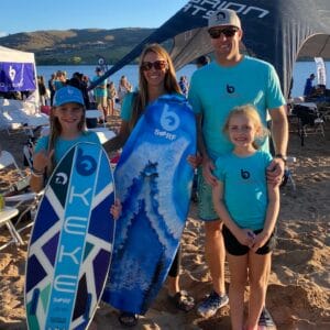 A family of four in matching blue outfits stands on the beach, looking like true Brabenders. The two children, proud Supreme Owners, hold surfboards as tents and chairs dot the background by the gently lapping waves.
