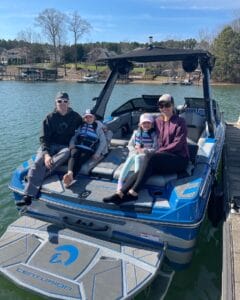 A family of four relaxes on a 2025 Centurion Ri230 boat docked at a picturesque lake, surrounded by lush trees and charming houses. The children, securely harnessed in life vests, are all smiles as they enjoy this Brabenders getaway.