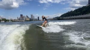 A person elegantly surfs a river beside the city skyline, with bridges, trees, and buildings forming a picturesque backdrop beneath a partly cloudy sky. In the distance, the sleek profile of the Centurion Ri230 adds a touch of modernity to this serene scene.
