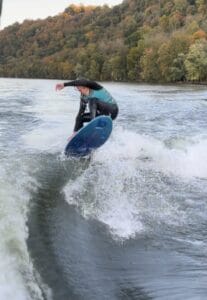 A person in a wetsuit is surfing on a river aboard a sleek Centurion Ri230, gracefully cutting through foamy waves, with a lush forested hill providing the perfect backdrop.