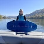 A person in a wetsuit stands on a Centurion Ri230, holding a blue surfboard emblazoned with "BLUSURF," against the serene backdrop of a calm river and tree-lined banks.