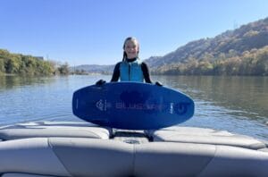 A person in a wetsuit stands on a Centurion Ri230, holding a blue surfboard emblazoned with "BLUSURF," against the serene backdrop of a calm river and tree-lined banks.
