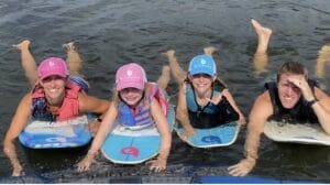 Four people in life vests and caps lie on paddleboards in the water, smiling and making hand gestures, as if celebrating their Supreme Owners' adventure.