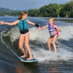 Two girls in life vests and swimsuits surf on a river wave from a sleek Centurion Ri230, extending their hands towards each other, surrounded by lush greenery and distant hills.