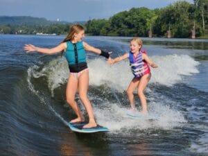 Two girls in life vests and swimsuits surf on a river wave from a sleek Centurion Ri230, extending their hands towards each other, surrounded by lush greenery and distant hills.