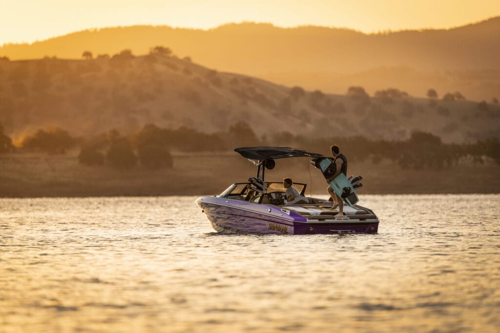 A Supreme S220 with several people onboard floats on a lake at sunset; one person stands holding a wakeboard while rolling hills rise in the background.