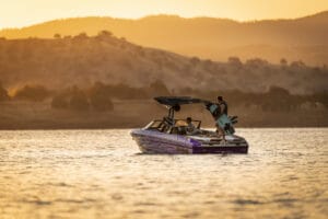 A Supreme S220 with several people onboard floats on a lake at sunset; one person stands holding a wakeboard while rolling hills rise in the background.