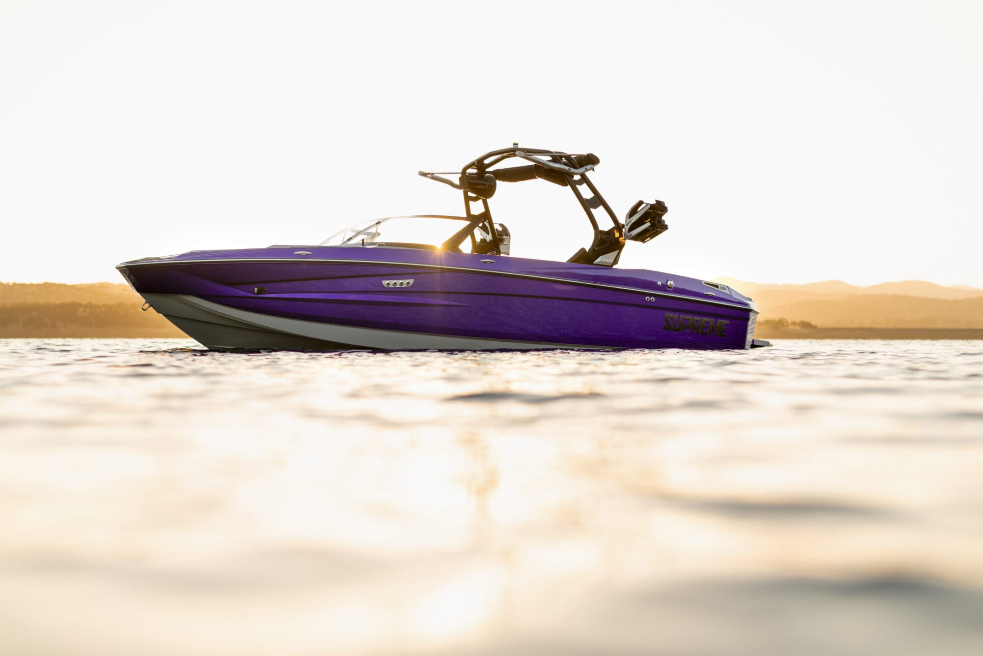A purple Supreme S220 speedboat with a wakeboard tower floats on calm water at sunset, with distant hills visible in the background.