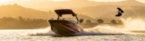 A Supreme Wake Boat glides across the lake at sunset, towing a wakeboarder mid-jump with majestic mountains in the background.