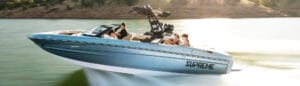 A blue and white Supreme wakeboat speeds across a sunny lake with several people seated inside, enjoying the ride.